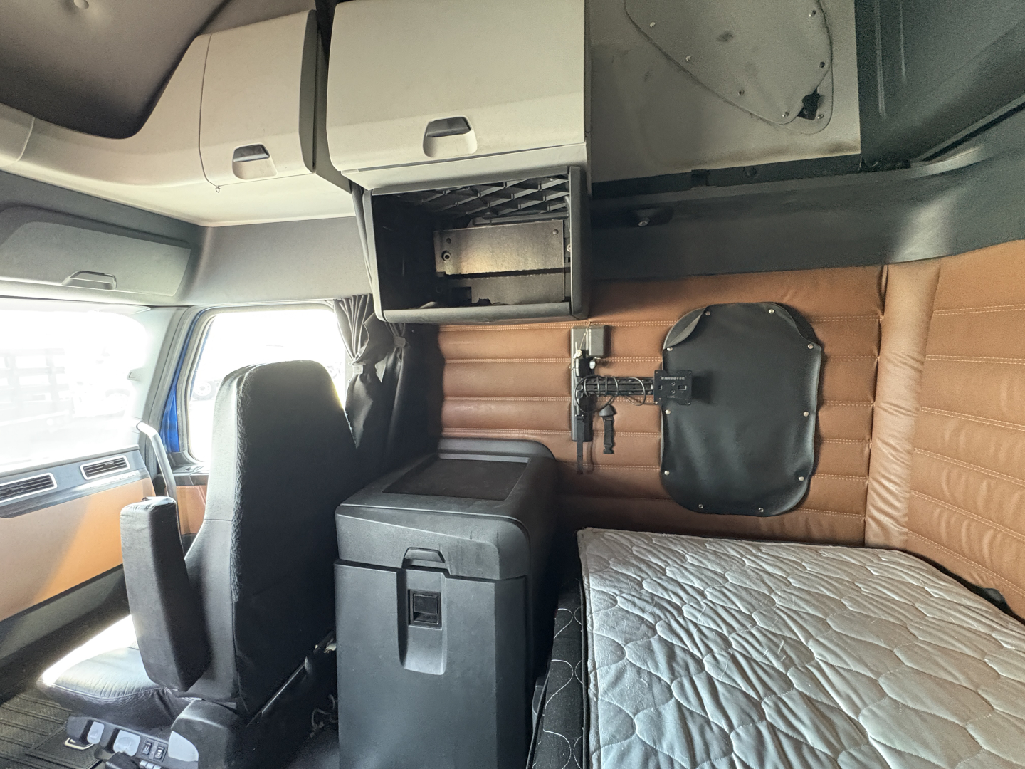 Cab interior of a Freightliner Cascadia 126 sleeper showing overhead storage, bunk area with mattress, and a mounted panel with latching hardware.