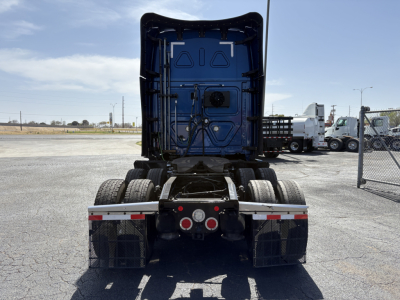 Rear view of a 2022 Freightliner Cascadia 126 tractor chassis showing twin rear axles and mud flaps, with blue sleeper cab door visible and a fenced lot background.