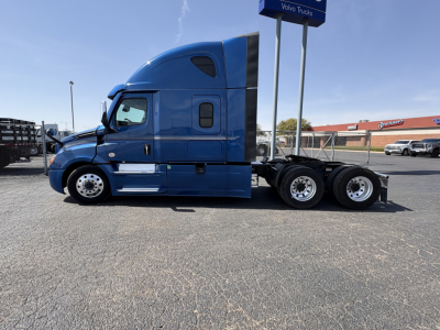 Cascadia 126 truck side profile in blue showing cab and rear twin-axle setup, no trailer, Freightliner branding on dealership sign in background