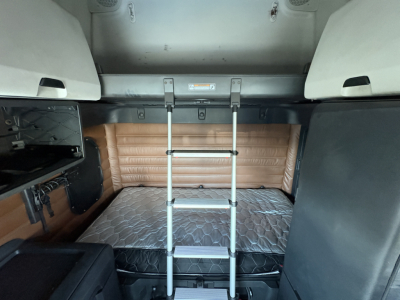 Cabin sleeper area of a Freightliner Cascadia 126 with a raised bunk ladder, tan padded wall, and a mattress in the sleeper compartment.