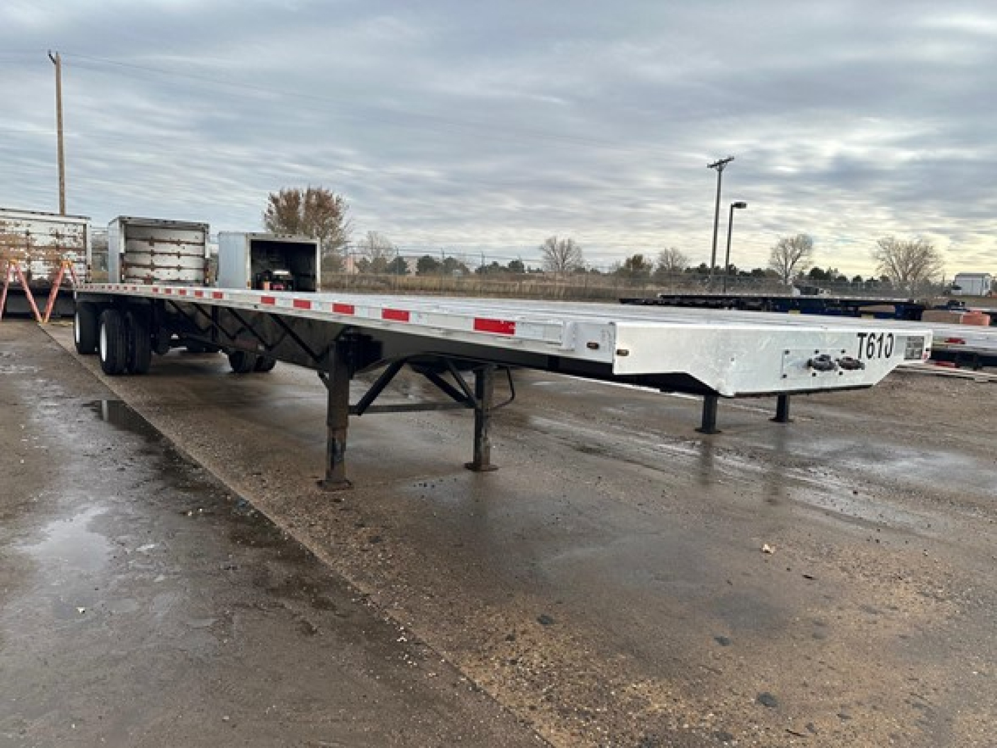 Flatbed trailer in a yard showing a long aluminum deck with support legs and tandem axles undercarriage.