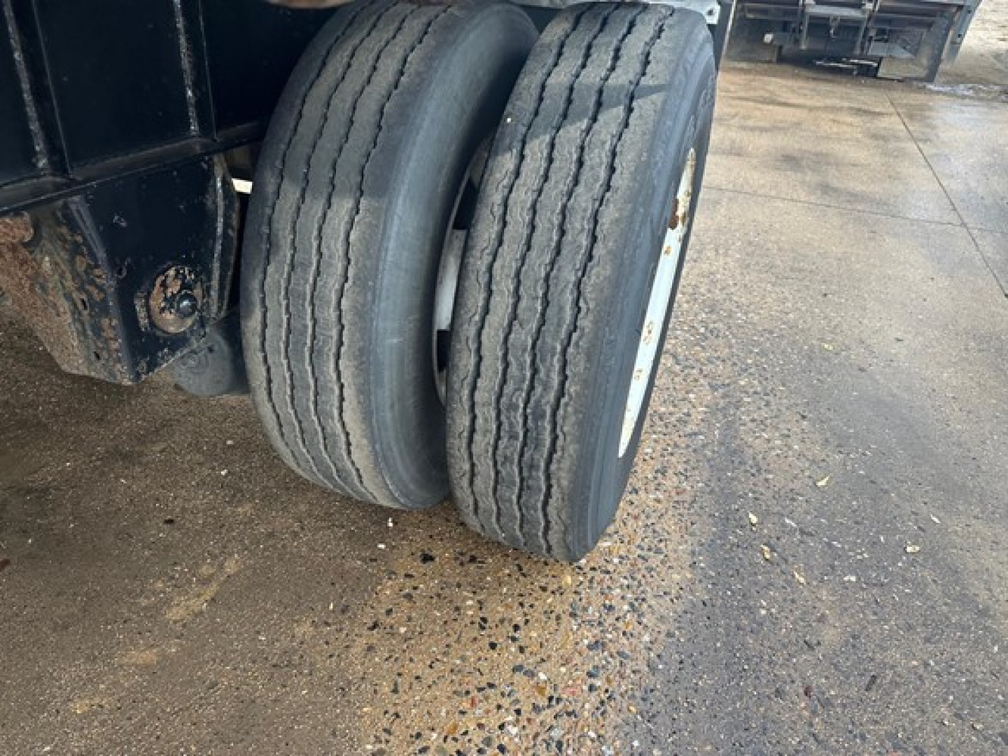 Trailer dual rear wheels with worn tires on a flatbed trailer. Visible are two wheels with white rims and part of the black frame; concrete floor and workshop background.