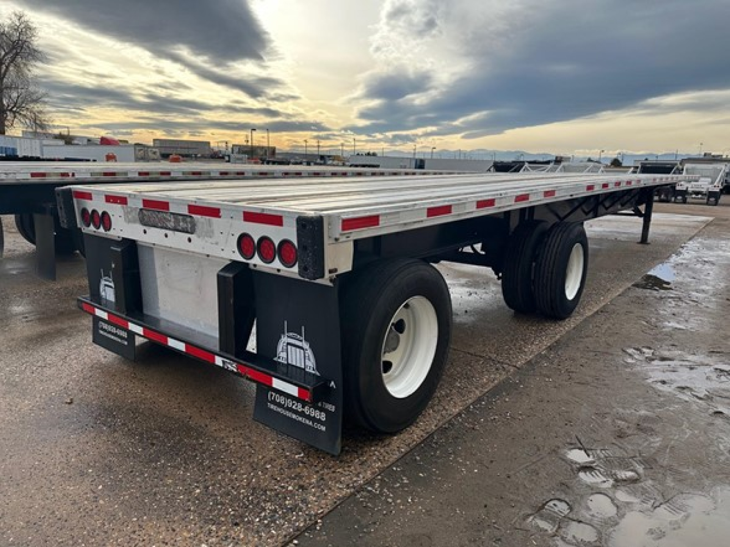 Flatbed trailer rear view showing Transcraft branding and dual rear mud flaps, with red and white reflective tape, dual rear tires, and license plate area.