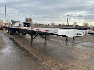 Flatbed trailer in a yard showing a long aluminum deck with support legs and tandem axles undercarriage.