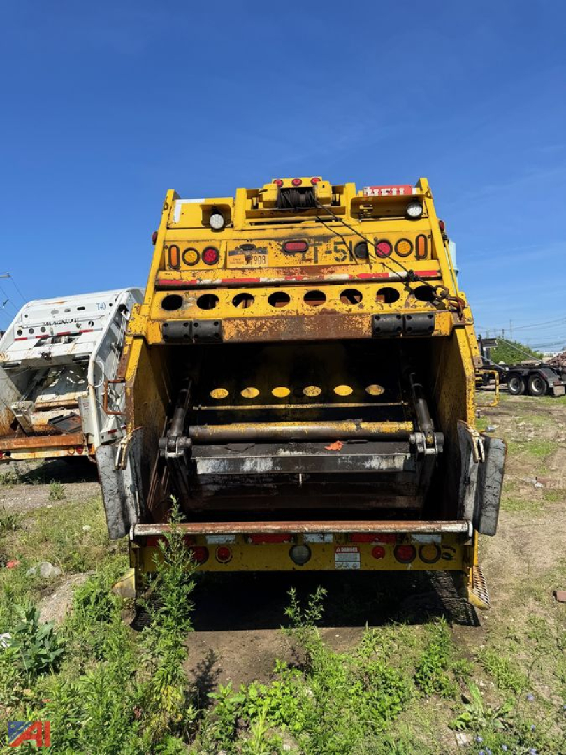 Yellow Mack garbage truck rear view showing compacting mechanism and tailgate with circular openings and warning lights.