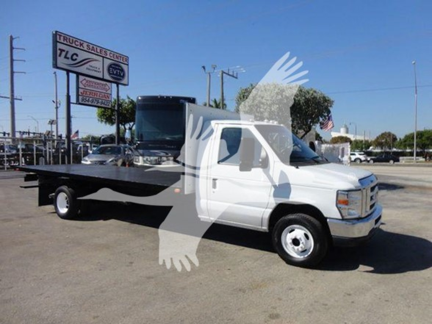 Flatbed tow truck in a dealership lot with Ford E-Series cab; visible text includes 'Truck Sales TLC' on a sign in the background and 'Ford' branding on the truck grille.