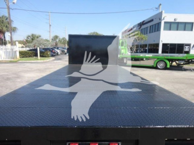 Flatbed truck bed surface with diamond plate texture and a faint graphic overlay, showing a landscape with a parked green and white bus in the background.