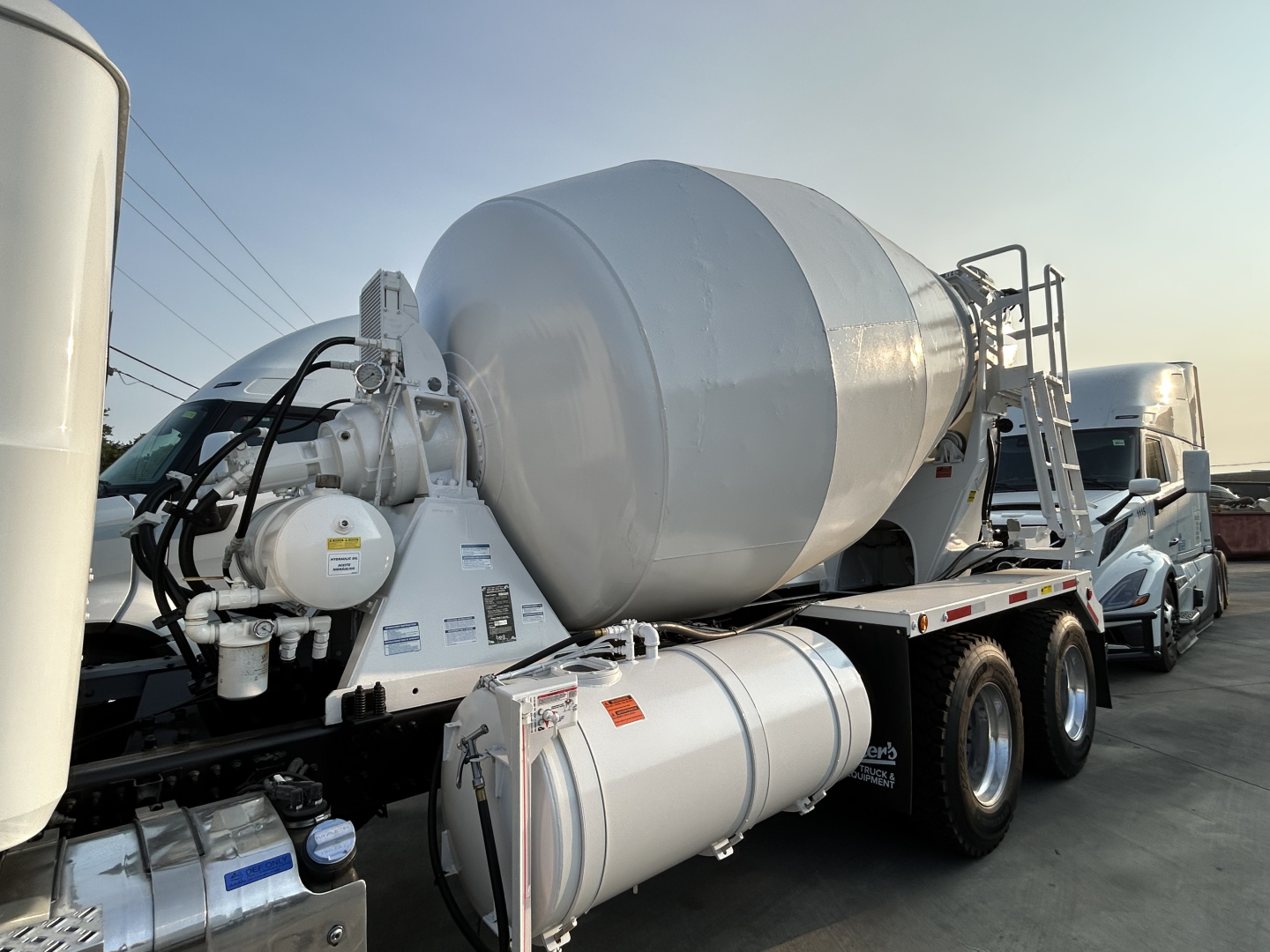 Mixer truck on a low-profile tilt with a gray cement mixer drum and auxiliary tanks, side view showing hydraulic pump and piping assembly.