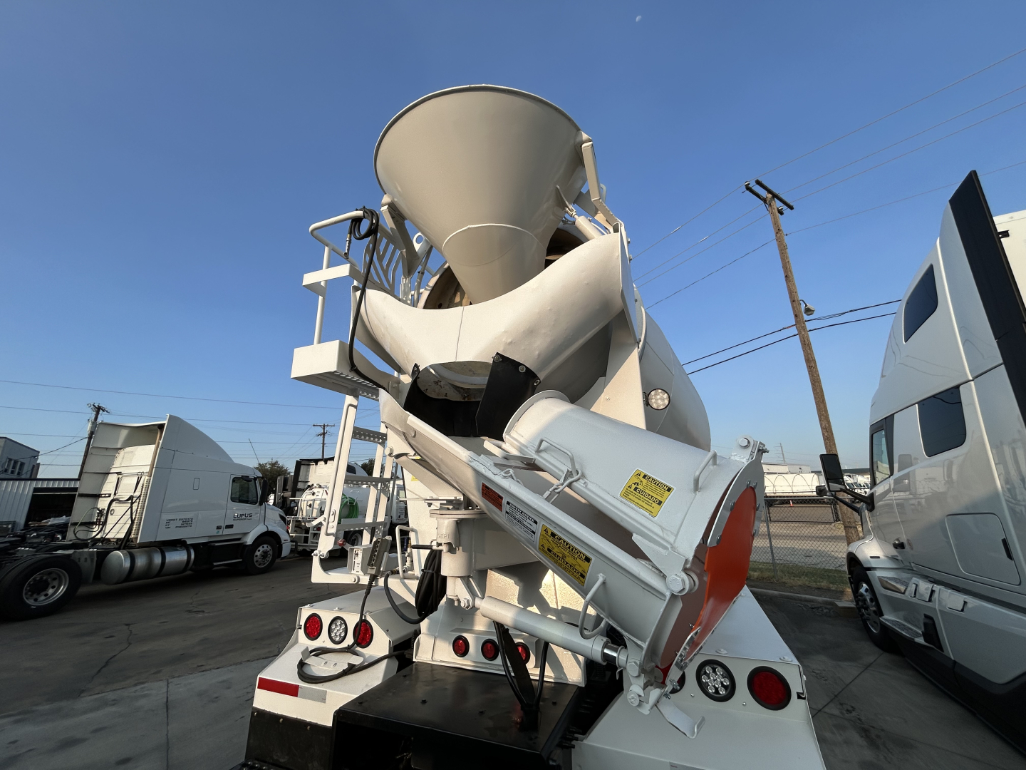 Mixer truck rear view with large concrete mixer drum and chute, white with red and yellow safety decals, visible text labels on chute and frame.