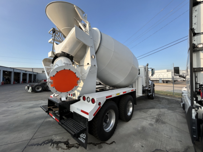 Mixer truck rear view showing concrete mixer drum and orange access hatch cap. dual rear axles with mud flaps and white cab visible.