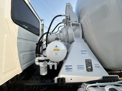 Mixer truck chassis showing hydraulic pump and control valve assembly near front wheel with warning labels and hoses