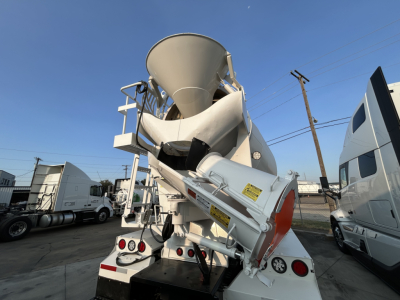 Mixer truck rear view with large concrete mixer drum and chute, white with red and yellow safety decals, visible text labels on chute and frame.