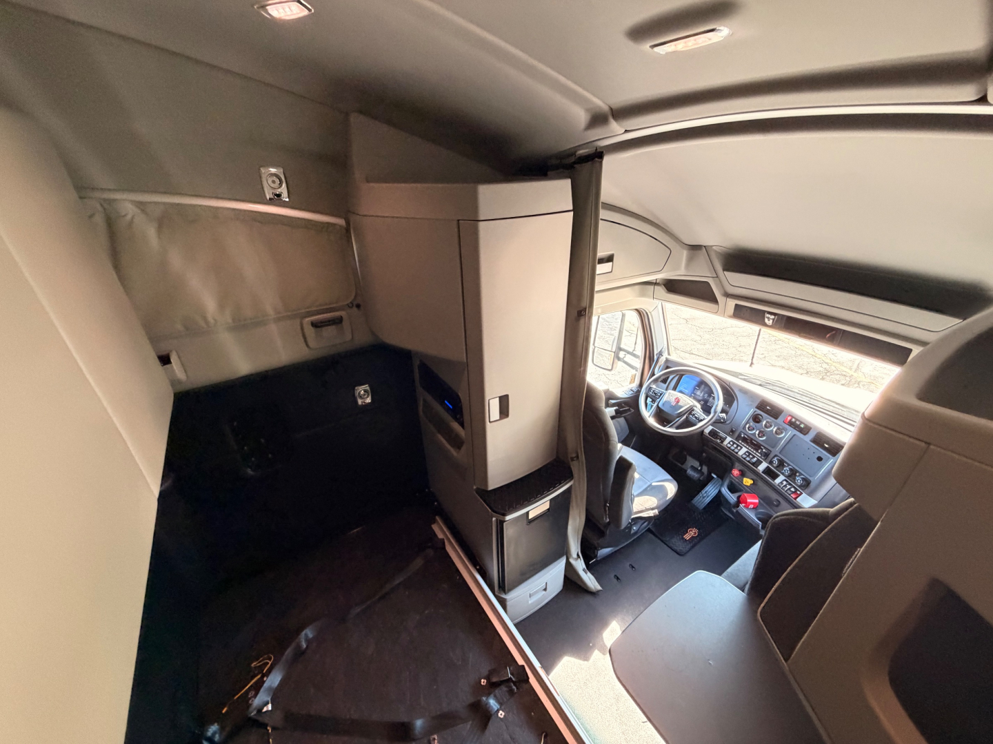 Cab interior of a Kenworth T680 tractor in cabin with sleeper area, showing dashboard, driver's seat, partition, and overhead storage.