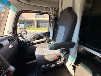Truck cab interior showing driver's seat and dashboard with controls; window and side mirror visible; curtain partition behind; appears to be a Kenworth T680 sleeper cab.