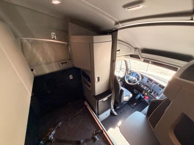 Cab interior of a Kenworth T680 tractor in cabin with sleeper area, showing dashboard, driver's seat, partition, and overhead storage.