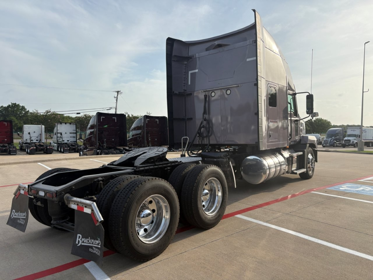 Truck rear/side view of a 2023 Mack AN64T70 conventional sleeper showing triple rear axles, fuel tanks, and cab with dark paint.