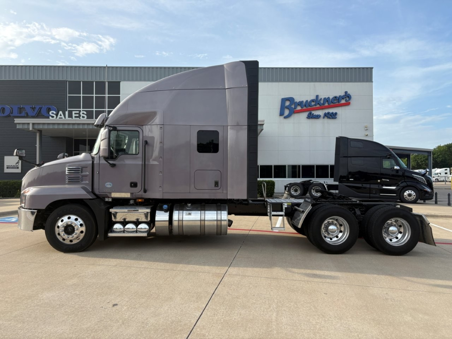 Mack AN64T70 conventional sleeper truck tractor side view with dual rear axles and gray cab, parked in lot in front of dealership building.