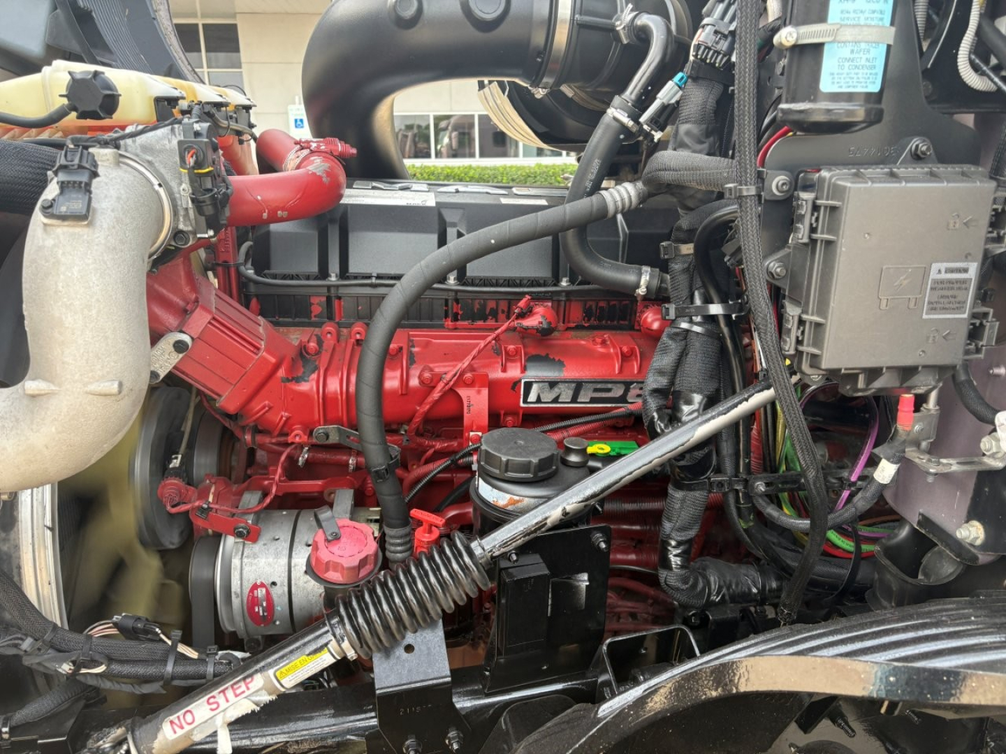 Mack AN64T70 engine bay showing red diesel engine, hoses, and components including coolant reservoir, belts, and wiring harness.