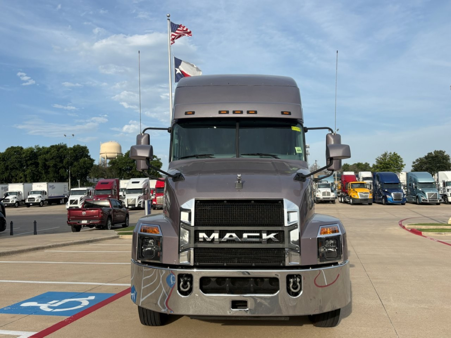 Truck front view of a 2023 Mack AN64T70 conventional sleeper showing large chrome grille with MACK letters, dual side mirrors, and surrounding dealership lot.