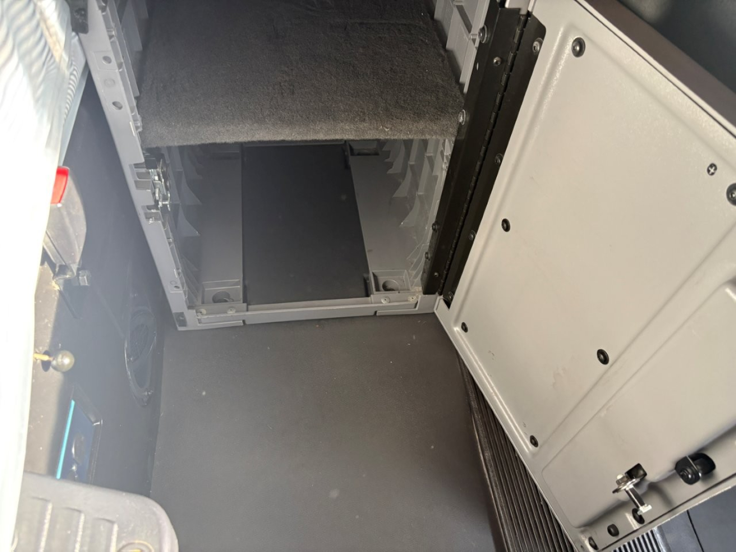 Truck cab interior showing storage compartments and metal framework under a carpeted floor; open panel with mechanical rails.