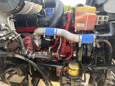 Engine bay of a 2023 Mack AN64T70 showing red engine block, blue silicone intercooler hoses, coolant reservoir, yellow oil filter, and various hoses and metal piping.