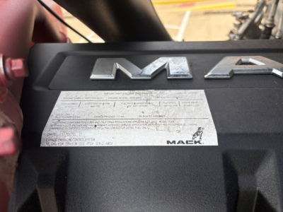 Mack engine bay with prominent 'MACK' emblem and a silver specification plate showing technical data; part of the engine cover is visible.