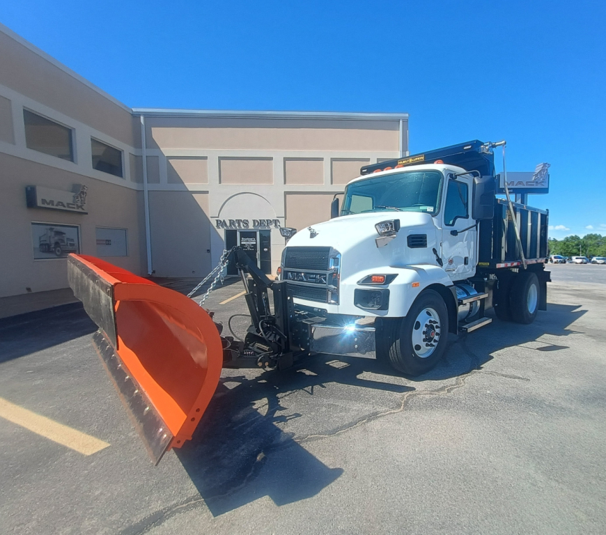 Front-left view of a white 2026 Mack MD7 plow truck with an orange plow attached, parked outside a service building.