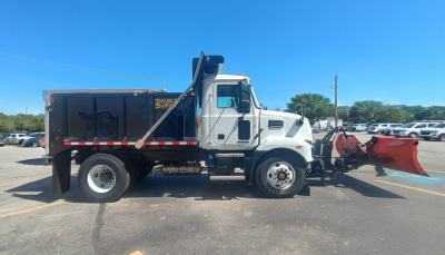 Truck side view of a 2026 Mack MD7 plow truck showing a large red snowplow at the front and a dumping body with lettering on the door. No visible labels legible besides storefront reflection.