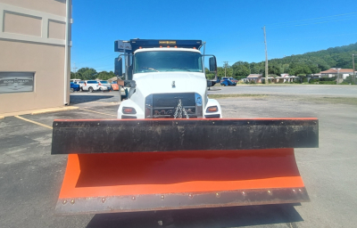 Front view of a white Mack MD7 plow truck with a large orange snow plow blade attached, visible grille, and windshield. No legible serials or stickers in view.