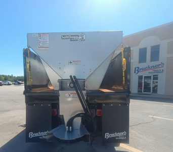 Rear view of a SaltDogg spreader mounted on a Mack MD7 truck chassis, showing stainless hopper, hydraulic hoses, and mount brackets; visible Buckner Truck Equipment logos.
