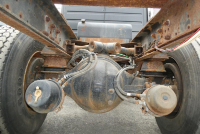 Truck rear axle and suspension view under Freightliner FL70 chassis showing differential housing, brake lines, axle components, and rusted frame rails.