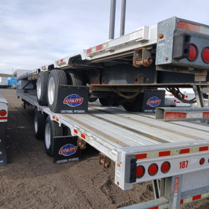Trailer showing a Manac drop deck trailer with dual rear axles, metal platform, side rails, and Utility Cheyenne Wyoming mud flaps; rear lights and reflective strips visible.