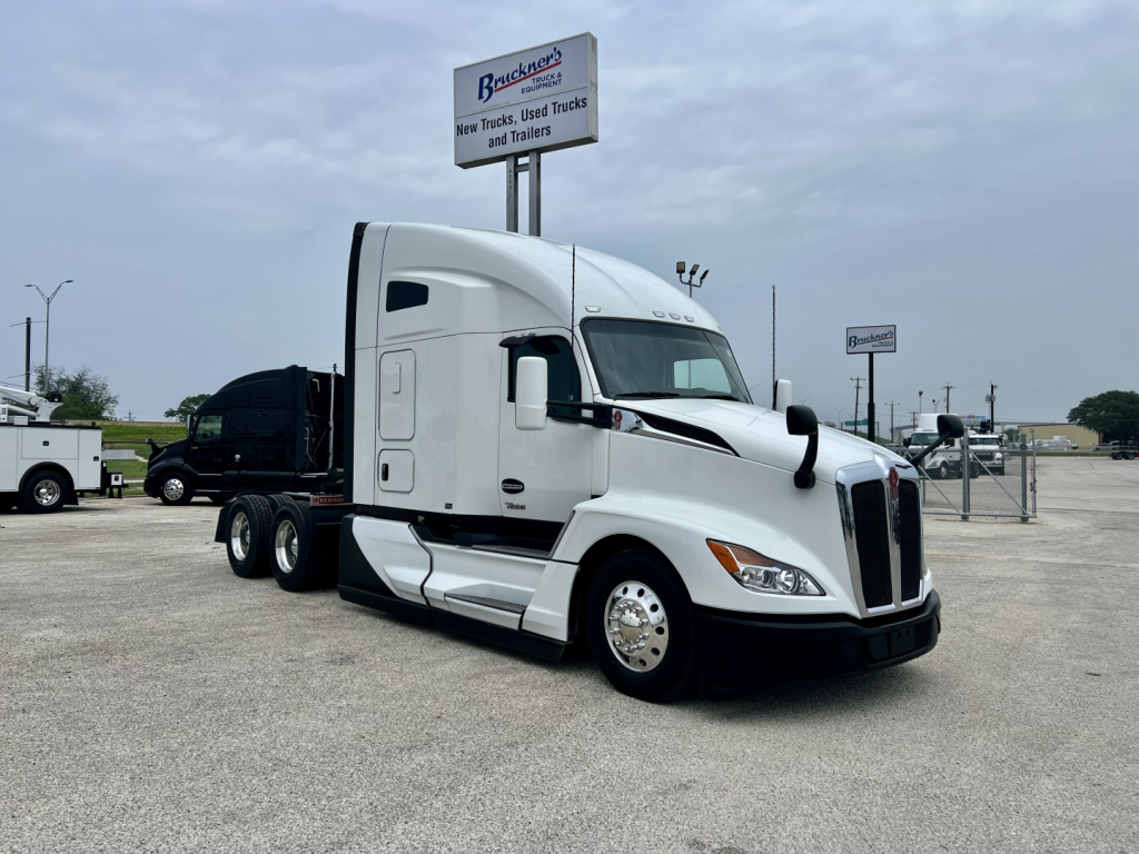 Kenworth T680 tractor-trailer in a dealership lot, white sleeper cab with aerodynamic hood and dual rear axles, front-view angle.