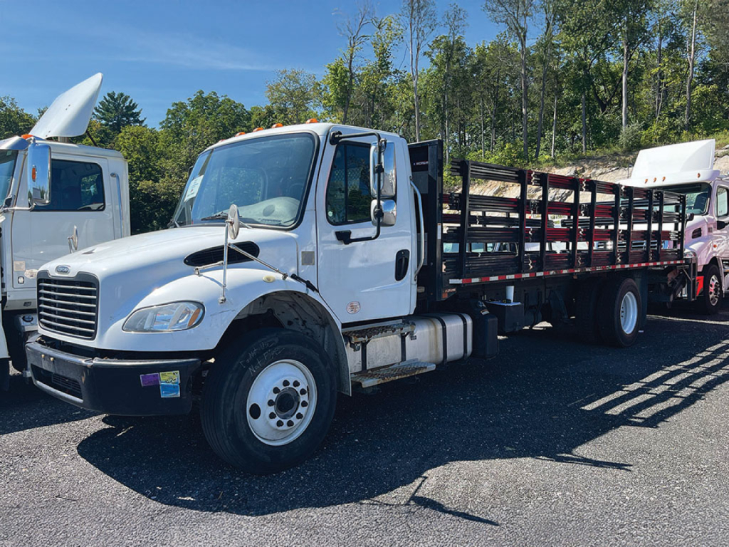 Truck with white cab and flatbed in a dealership lot showing Freightliner M2 106 Business Class style. Visible text on cab and decals, front grille, and side mirrors.
