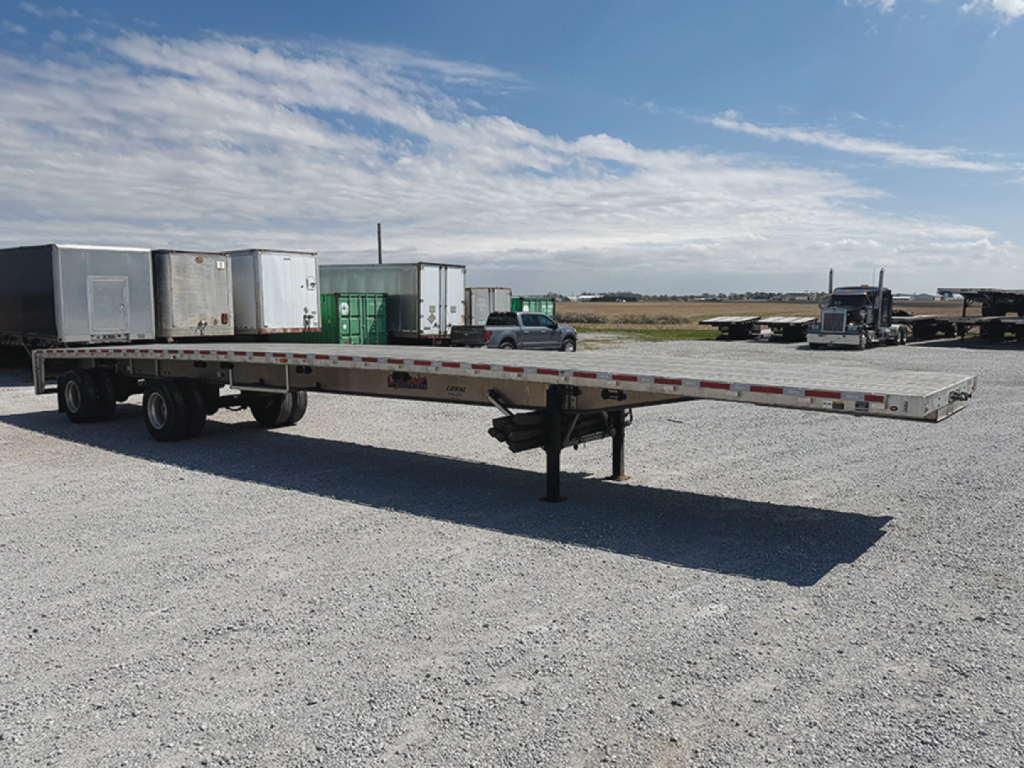 Flatbed trailer in an open lot showing a long aluminum deck and drop-leg jack.