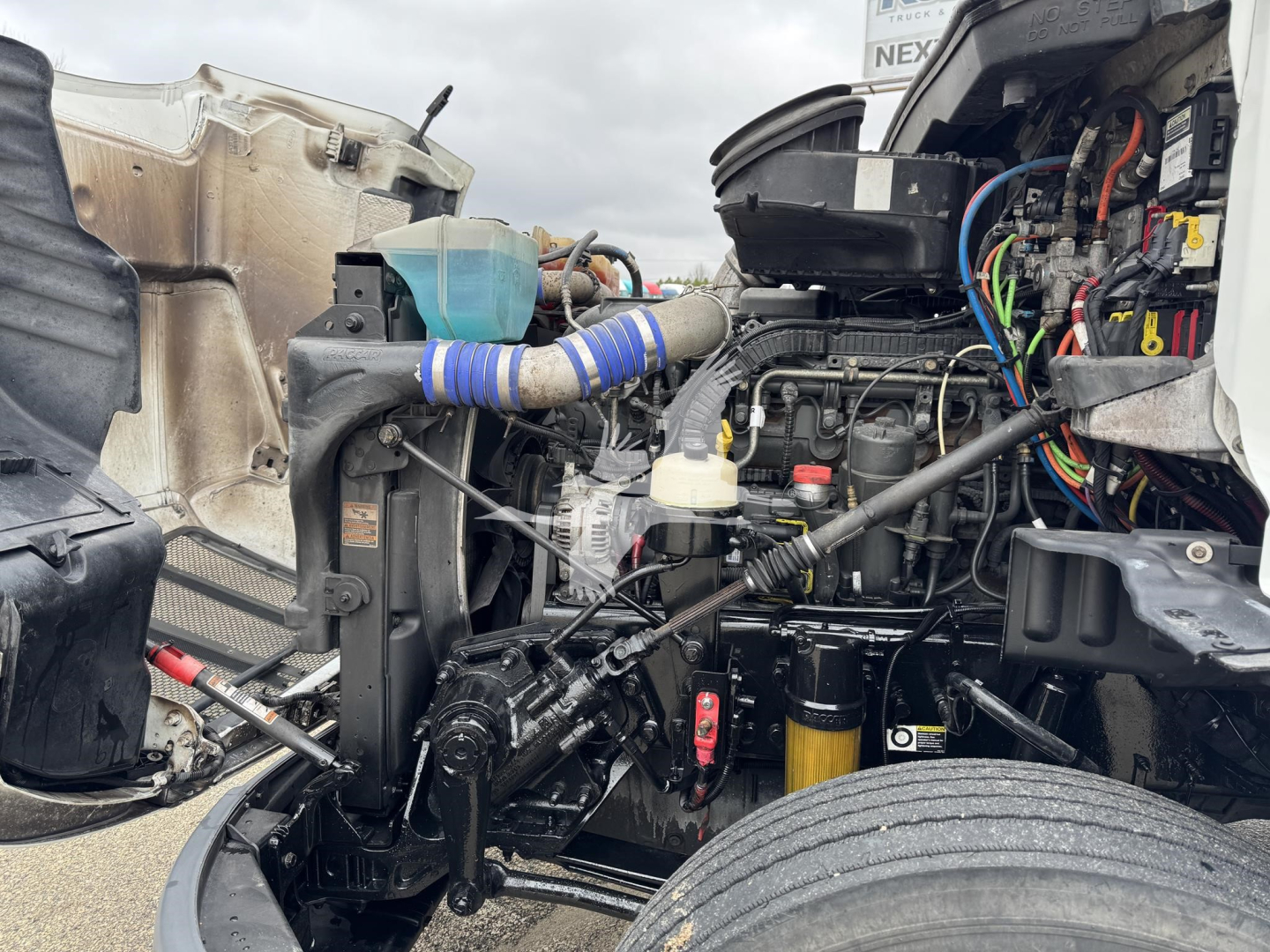 Peterbilt 579 engine bay view with exposed components including wiring harnesses, hoses, and hydraulic lines; visible belt tensioner and air intake; engine block in view.