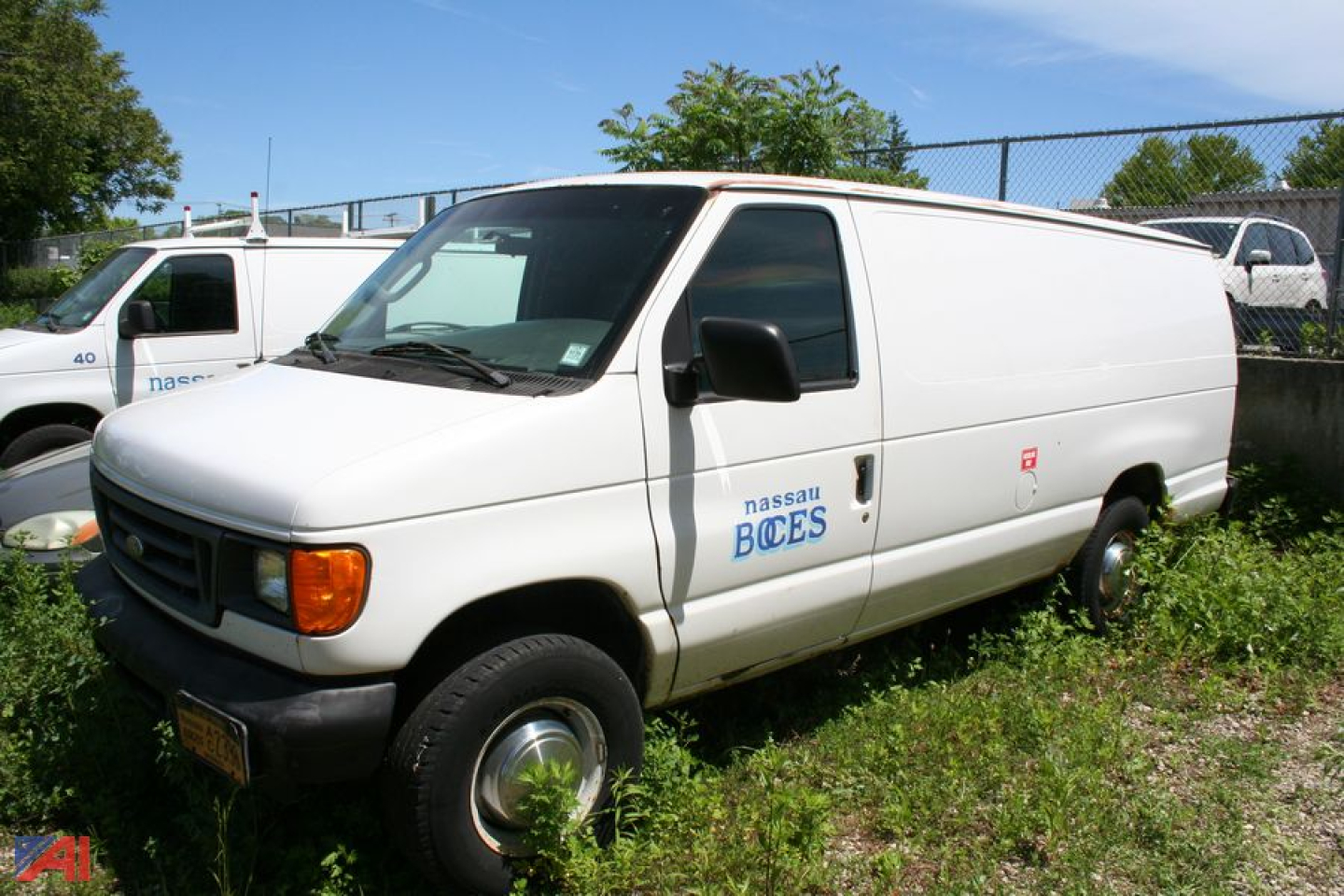 White 2006 Ford E350 cargo van with Nissan BOCES logo on side, black front bumper, sliding side door, parked on grass with chain-link fence background.