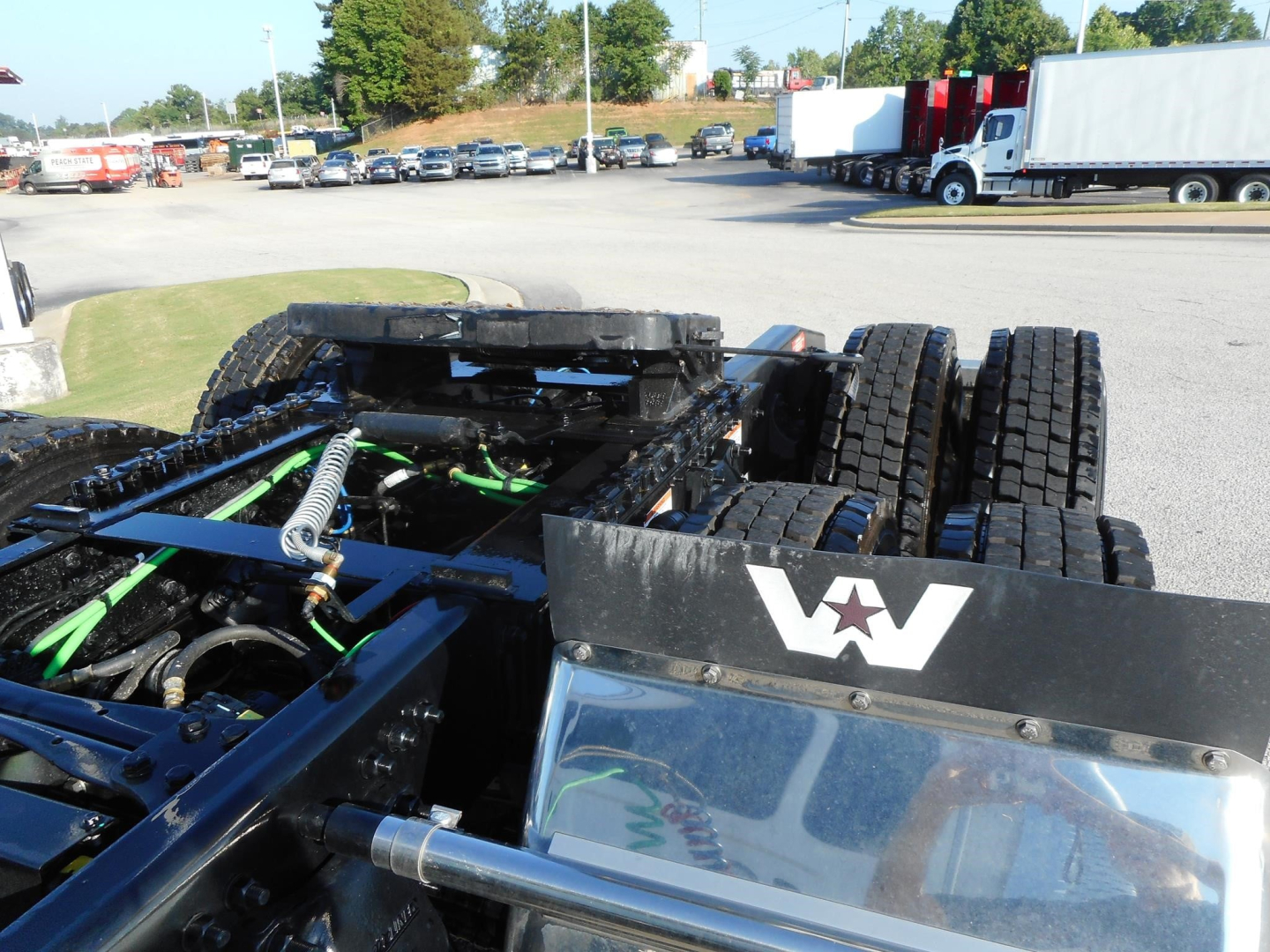 Western Star 49X truck chassis with dual rear axles and green structural components visible; emblem 'W' on black metal frame near cab; multiple trucks in background.