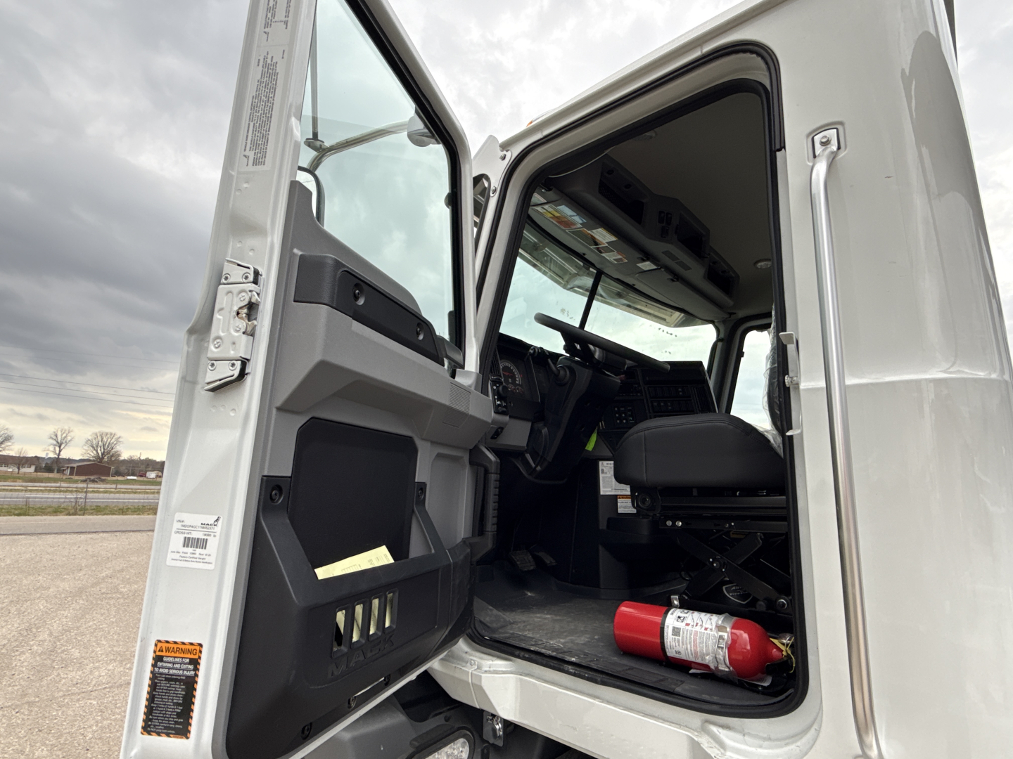 Trucks cab interior view of a 2026 Mack GR64B roll-off truck showing open door and driver seat, with a fire extinguisher on the floor. VIN 1M2GR4GC1TM052371 and stock 69806 mention on image.