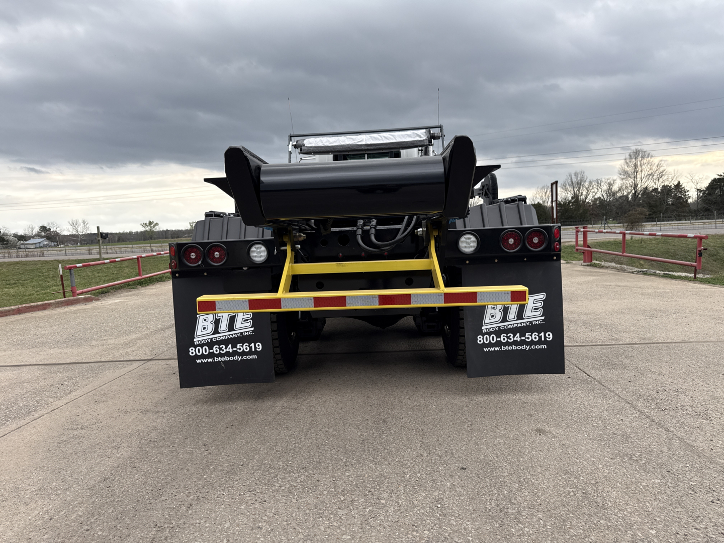 Back view of a new 2026 Mack GR64B roll-off truck showing rear hitch, hydraulic arms, and mud flaps with dealership contact numbers.
