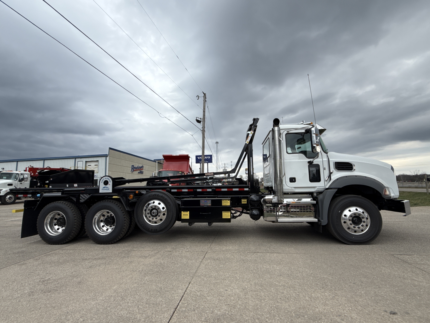 2026 Mack GR64B roll-off truck side view showing cab, long chassis, wheel configuration with six rear wheels, loading arms, and hydraulic components.