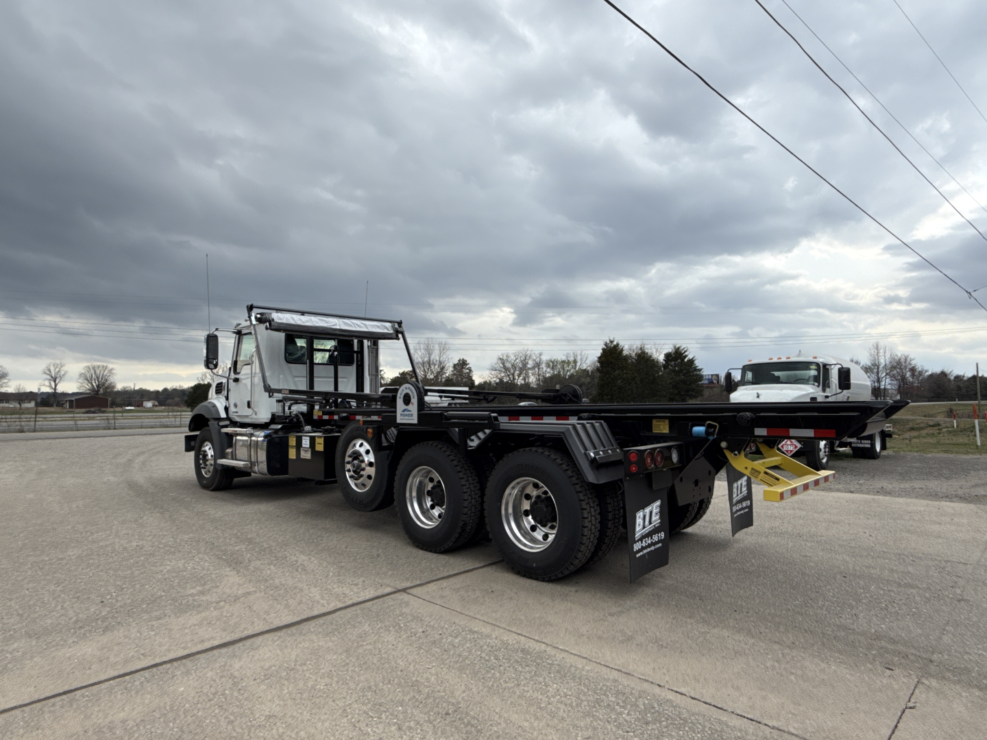 Mack GR64B roll-off truck with long wheelbase, three-axle configuration, flatbed/tipping unit, and a yellow ramp/hook at the rear. Cab is white with black chassis and dual rear wheels.