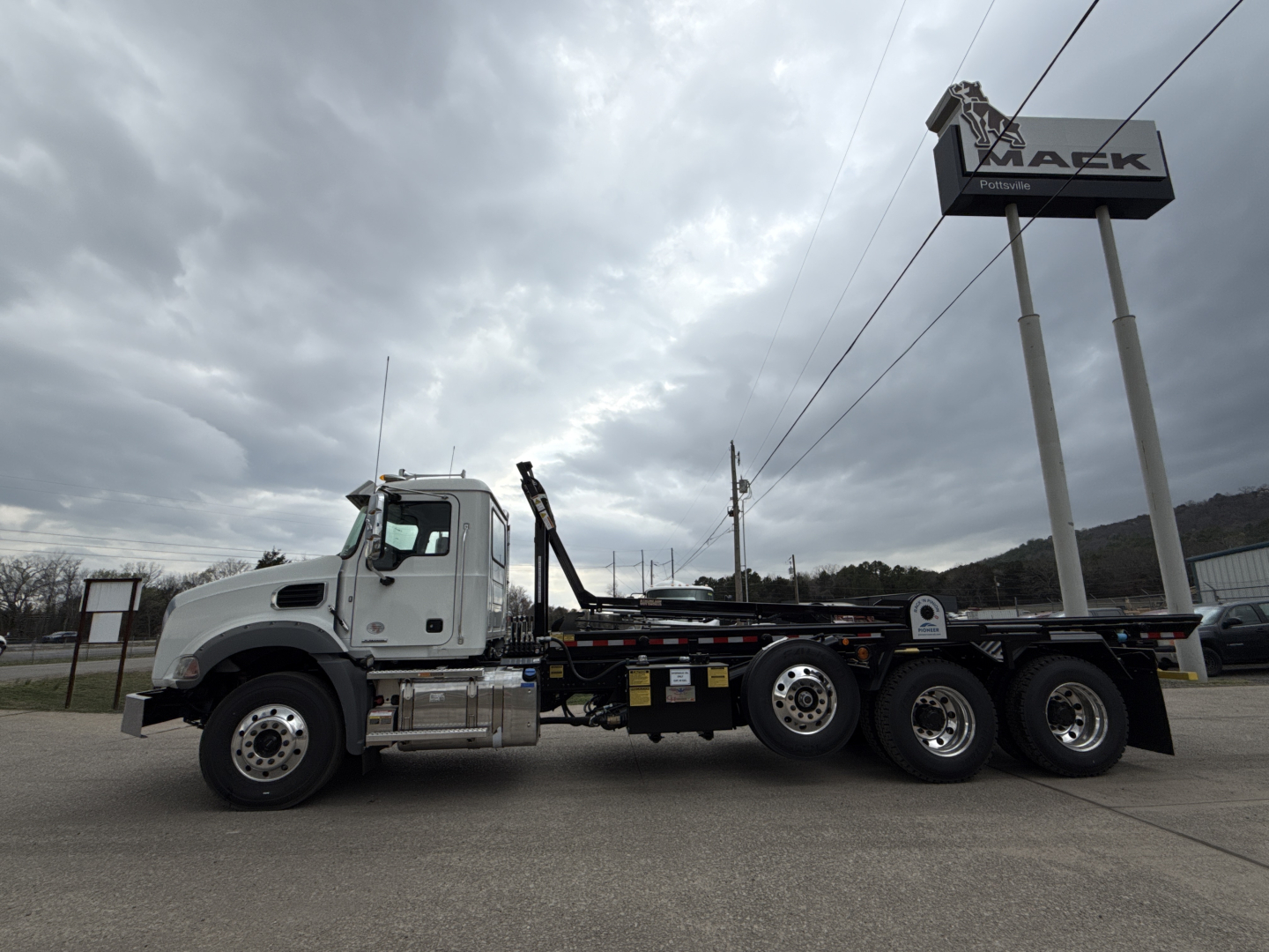 Mack GR64B rolloff truck side view showing cab, chassis, and rear axles with towing equipment; dealership sign in background.