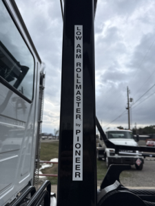 Vertical label on a Mack GR64B roll-off truck showing 'LOW ARM ROLLMASTER by PIONEER' along the structural pillar in an outdoor lot.