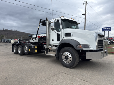 Mack GR64B rolloff truck side view showing white cab, three axles, and flatbed with crane/oil rack. VIN 1M2GR4GC1TM052371 visible on listing.