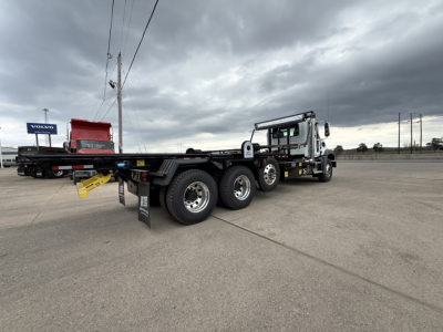 Mack GR64B roll-off truck with extended container bed parked in lot; visible text includes Mack logo on cab, DOT license plates, and a Volvo sign in background.