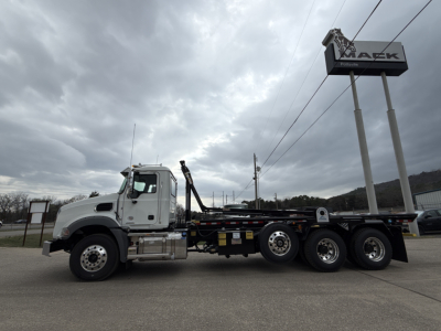 Mack GR64B rolloff truck side view showing cab, chassis, and rear axles with towing equipment; dealership sign in background.