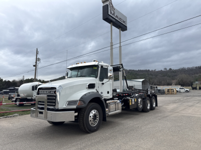 Mack GR64B truck in a lot showing front cab and 6x4 rear axles with roll-off body; visible text on side and sign in background.