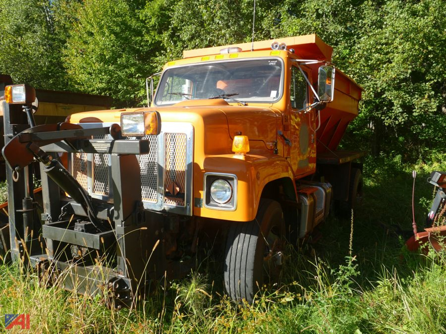 Industrial truck with orange stone spreader body, front grille, and snowplow attachment, viewed from left-front angle in grassy lot.