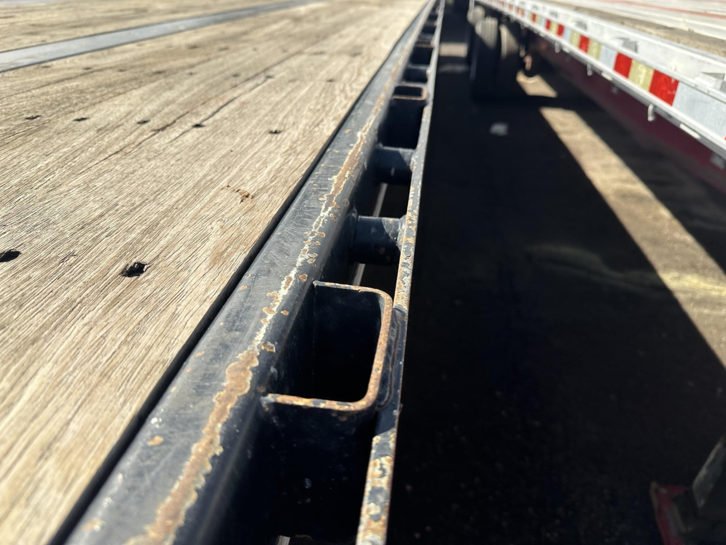 Flatbed trailer deck edge with stake pockets and wooden plank surface; side rails show wear and rust spots along the metal beam.